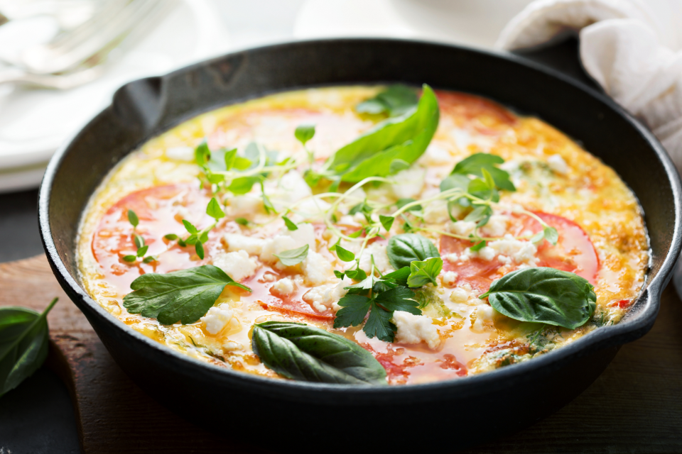 Vegetable frittata topped with cherry tomatoes, herbs, and melted cheese cooking in a black iron cast pan